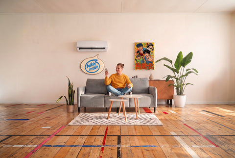 A man wearing a yellow knitted jumper is sitting crossed-legged on a grey couch. He holds a smart phone in his left hand and he is smiling, on the wall behind him is an Air Conditioning unit with a speech bubble that says "Faikin inside". There is a manga poster on the wall that says "automate".