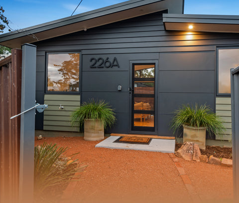 A small house can be seen through an open gate. The house is grey in colour and the house numbers 226A are on the wall next to the front door. There are two pot plants either side of the door. The welcome mat says "Hello".